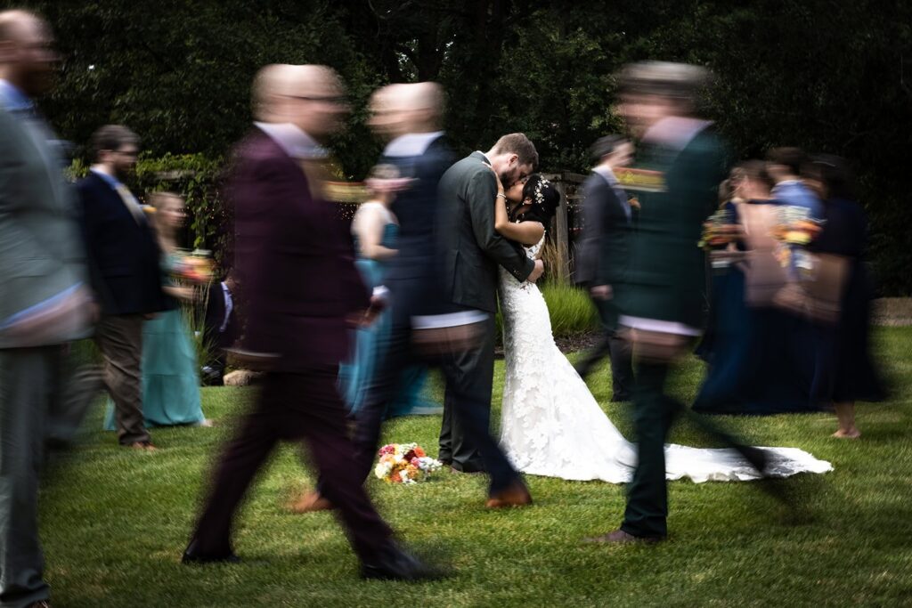 Artistic portrait of bride and groom kissing while guests walk around them